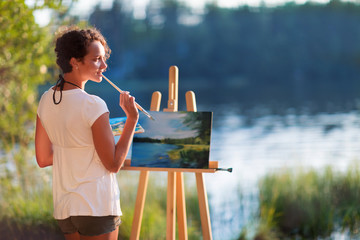 Fototapeta premium Young pretty woman artist draws paints a picture of a lake on open plain air outdoors