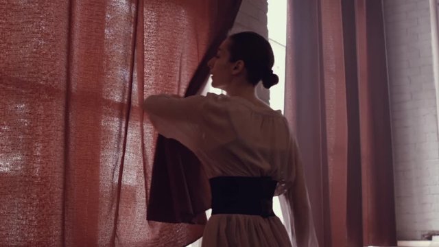 Girl approaches large panoramic window and shades it. Young girl with  beautiful dress from the back in the room next to  window. Girl ballerina in big studio room. Plot for the ballet.