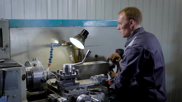 Young industrial worker standing behind lathe machine indoor, working with metal pieces.