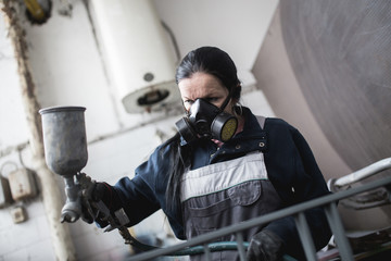 Strong and worthy woman doing hard job. She using industrial spray compressor for painting some metal products.