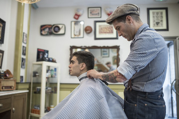 Barbero moderno con vestimenta vintage corta el pelo a joven muchacho veiteañero en barbería.