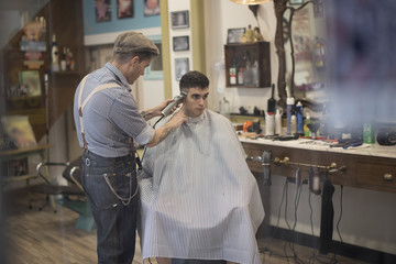 Barbero moderno con vestimenta vintage corta el pelo a joven muchacho veiteañero en barbería.