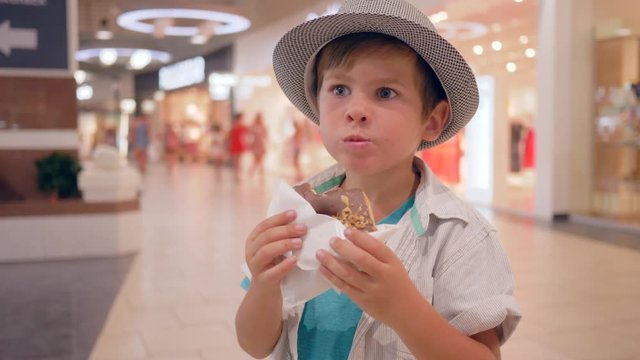 Children food, cute happy kid into Hat eating chocolate doughnut in shopping mall close-up