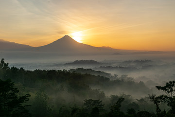 Scenic view of sunrise behind Merapi volcano, misty jungle and Borobudur temple in Indonesia 