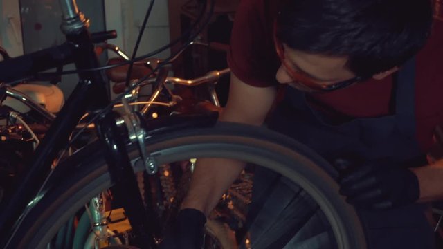 theme small business bike repair. Young brunette Caucasian man in protective glasses, gloves and fartuhe uses hand tools to repair and tuning Rim Brakes and wheel spinning bike in a garage workshop