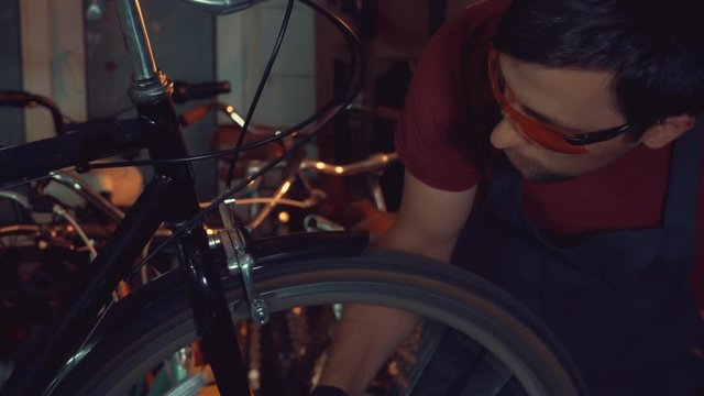 theme small business bike repair. Young brunette Caucasian man in protective glasses, gloves and fartuhe uses hand tools to repair and tuning Rim Brakes and wheel spinning bike in a garage workshop