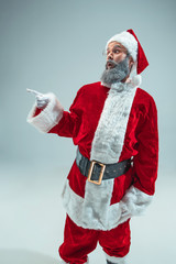 Fototapeta na wymiar Funny guy with christmas hat posing at studio pointing to left. New Year Holiday. Christmas, x-mas, winter, gifts concept. Man wearing Santa Claus costume on gray. Copy space. Winter sales.