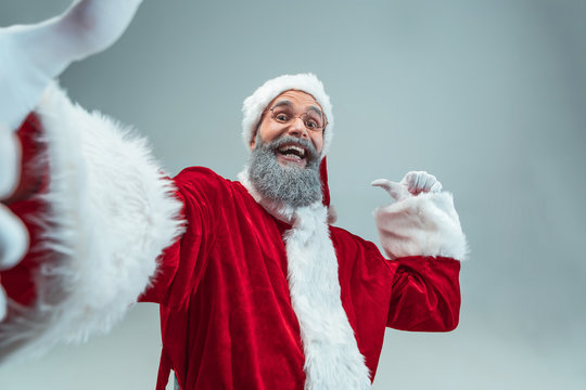 Funny guy with christmas hat posing at studio pointing to left. New Year Holiday. Christmas, x-mas, winter, gifts concept. Man wearing Santa Claus costume on gray. Copy space. Winter sales.