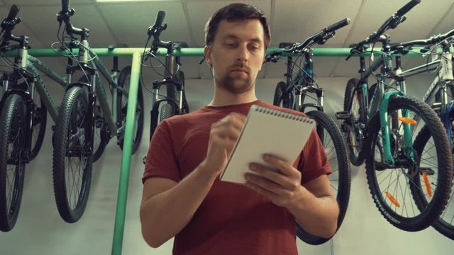 theme of small business selling bicycles. Young Caucasian male brunette small business owner, store manager uses notepad and pen makes notes, checklist at bicycle store