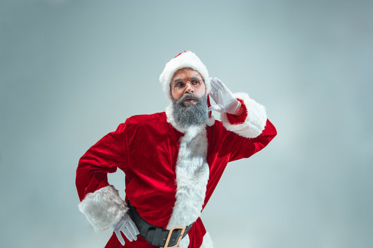 Funny eavesdropping guy with christmas hat posing at studio. New Year Holiday. Christmas, x-mas, winter, gifts concept. Man wearing Santa Claus costume on gray. Copy space. Winter sales.