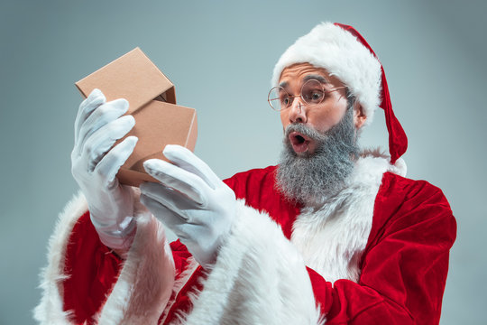 Funny guy with christmas hat posing at studio with gift. New Year Holiday. Christmas, x-mas, winter, gifts concept. Man wearing Santa Claus costume on gray. Copy space. Winter sales.