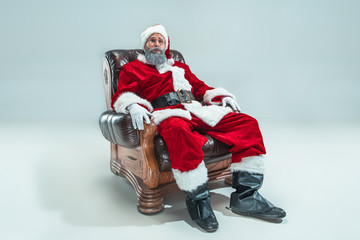 Fototapeta na wymiar Funny serious guy with christmas hat sitting at studio. New Year Holiday. Christmas, x-mas, winter, gifts concept. Man wearing Santa Claus costume on gray. Copy space. Winter sales.