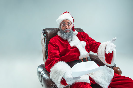 Funny serious guy with christmas hat sitting at studio with a letter of desire. New Year Holiday. Christmas, x-mas, winter, gifts concept. Man wearing Santa Claus costume on gray. Copy space. Winter