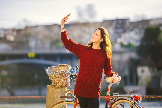 A beautiful young woman with a retro red bicycle is making a photo of herself in the old city of Europe on the River Rhine embankment in the Swiss city of Basel. Sunny warm day in winter