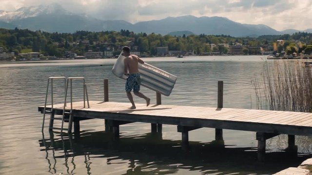Slow-motion man jumping from a dock on a mattress in Worthersee lake, Austria mountain view sunny day tracking shot 