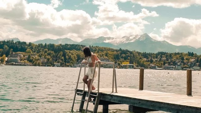  Young woman in swimwear climbs down the stars on a dock in Worthersee lake, mountain view windy hair in slow-motion