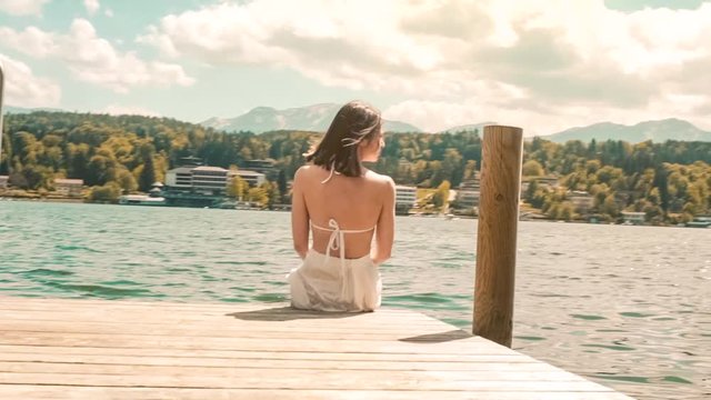 Young woman in swimwear sitting and admiring the mountain view on a dock in Worthersee lake, Austria windy hair in slow-motion