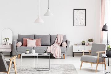 Armchairs and tables in white living room interior with poster and pink blanket on grey sofa. Real photo