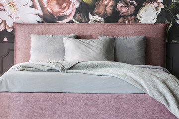 Grey blanket and cushions on pink bed in feminine bedroom interior with flowers wallpaper. Real...