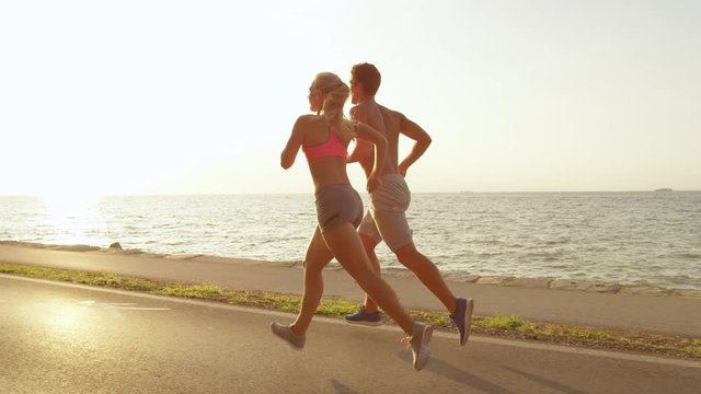 SLOW MOTION, LENS FLARE: Happy young man and woman jogging along the beautiful coastal road in Slovenia Cheerful Caucasian fit couple enjoying a relaxing run in the summer sun by picturesque seaside.