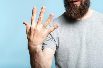 bearded man showing number five with his hand. cropped shot of a male torso on blue background. casual hipster in grey t-shirt counting gesture.