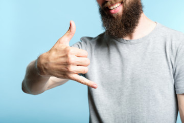 bearded man showing phone call gesture or shaka sign with hand. cropped shot of a male torso on blue background. casual hipster in grey t-shirt.