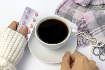 Hands in white sweater holding a blister of pills with white ceramic cup of coffee and pink knitted plaid lying near on white table
