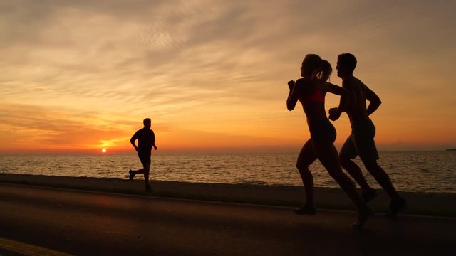 SLOW MOTION, COPY SPACE, SILHOUETTE: Fit young couple enjoying a beautiful summer evening by jogging by the ocean. Athletic man and woman running together down the scenic coastal pedestrian trail.