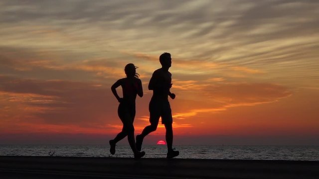 SLOW MOTION, SILHOUETTE: Athletic young man and woman run over the red sun setting behind the ocean. Cinematic shot of fit couple jogging shirtless along the ocean on a picturesque summer evening.