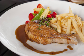 Grilled pork chop steak and vegetable salad, french fries with brown sauce in a white plate on tho wooden table background