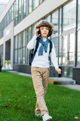 full length view of boy with backpack talking by smartphone and looking at camera