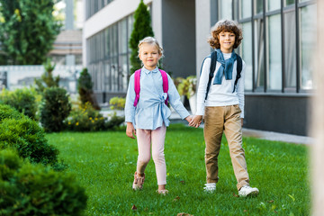 adorable schoolkids with backpacks holding hands and walking on green lawn