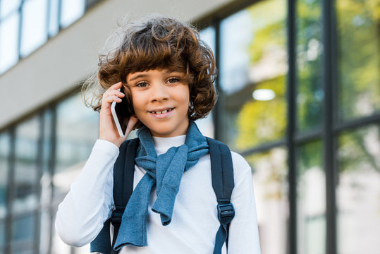 cute preteen schoolboy with backpack talking by smartphone and smiling at camera