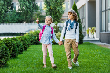 cute little schoolkids with backpacks holding hands and looking away while walking on green lawn