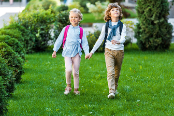 Fototapeta premium full length view of adorable happy schoolkids with backpacks holding hands and walking on green lawn