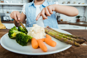 cropped shot of child holding fork with knife and eating healthy vegetables