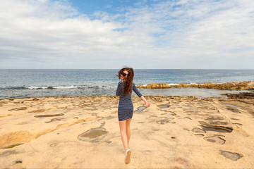 Fototapeta na wymiar curly-haired girl in a striped dress and sneakers gaily runs along the lava beach of the ocean sea shore on a sunny joyful day