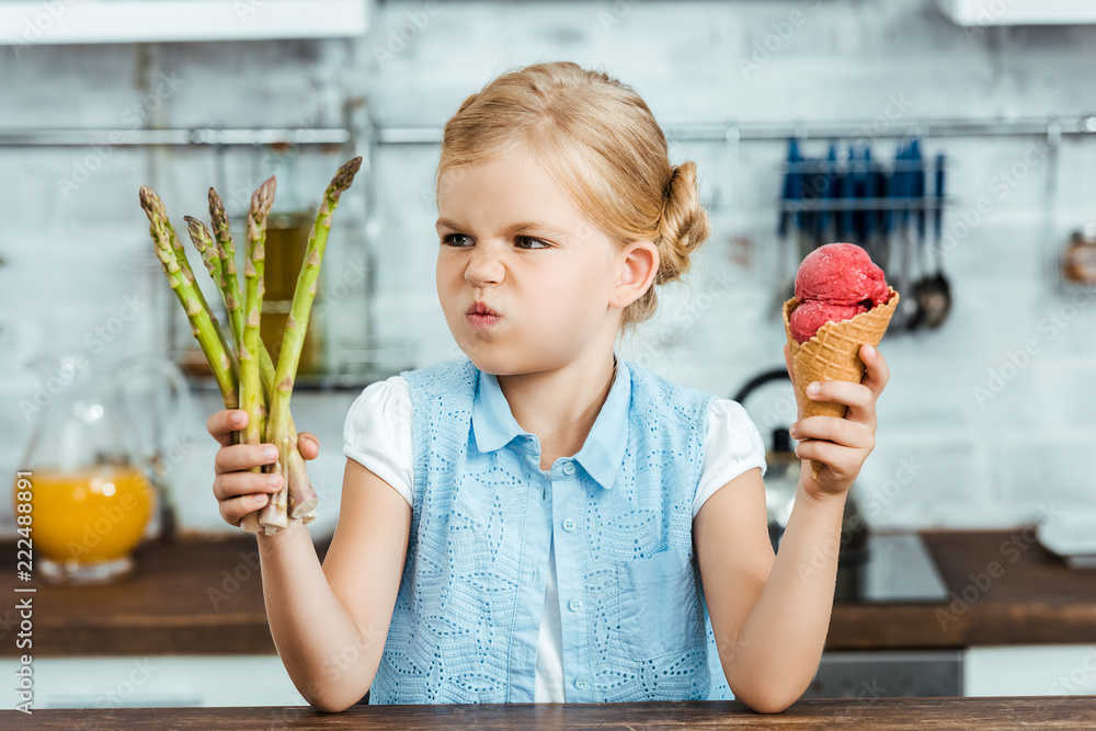 Wall mural unhappy child holding delicious ice cream cone and healthy asparagus