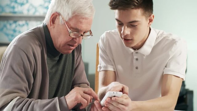 Medium shot of young volunteer teaching elderly man in glasses how to use smartphone and pay bills online using application