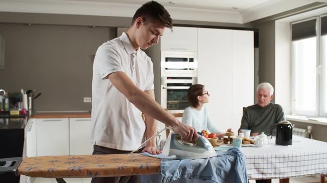Tracking shot of young male volunteer ironing clothes while his female friend sitting at table in the background and talking to retired man in kitchen