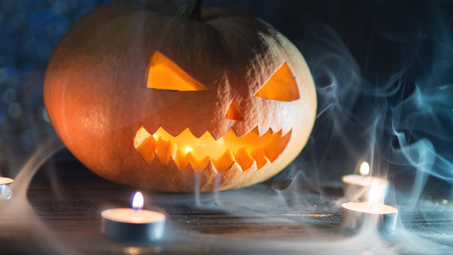 Orange crazy pumpkin lantern looks through the smoke. Dark background with candles