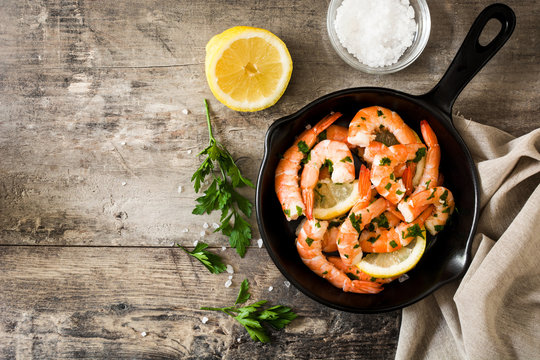 Grilled shrimps, parsley and lemon in iron pan on wooden table. Top view. Copyspace