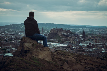 Top view of Edinburgh Castle, town. In the foreground, a man is sitting with his back looking at the city, Edinburgh, Scotland, United Kingdom