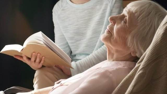 Tilt up shot of elderly lady relaxing in rocking chair and listening to young female caregiver who is reading book aloud to her in the background
