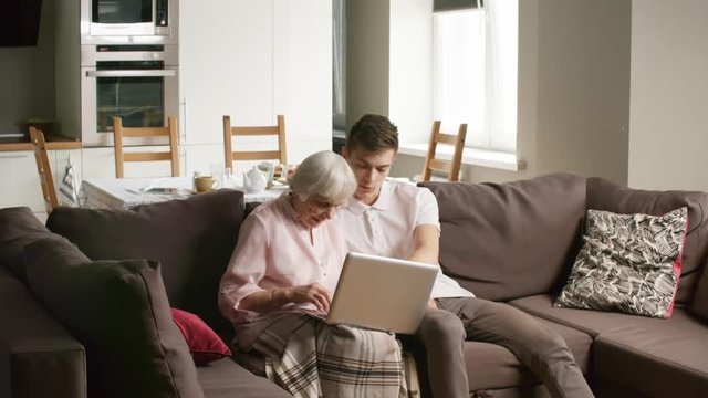 Interested retired woman and her grandson or volunteer sitting on sofa in living room and using laptop computer