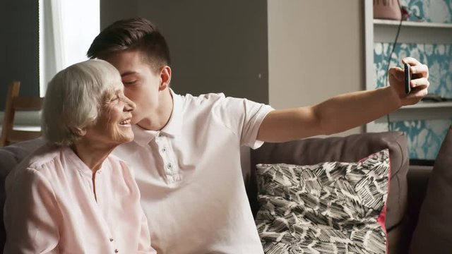 Medium shot of young man sitting on sofa next to grandmother and kissing her on the cheek while taking selfie with smartphone together