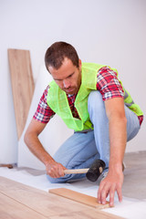 Young man wearing reflective safety vest installing laminate flooring in new apartment or house, holding a hammer. Home improvement and renovation concept