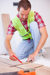Young man wearing reflective safety vest installing laminate flooring in new apartment or house, measuring and marking tiles for cutting. Home improvement and renovation concept