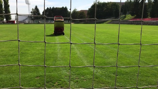 Grass-cutting vehicle cuts grass on football field, lines after grass-mower on green grass, view from goal net to playing field