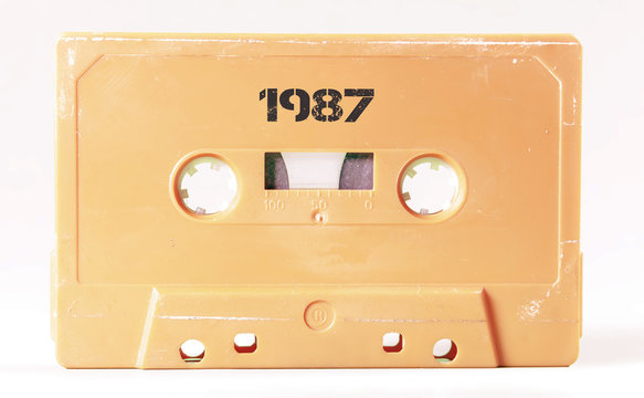 A vintage cassette tape from the 1980s era (obsolete music technology) with the text 1987 printed over it, stencil font. Color: cream, sand. White background.
